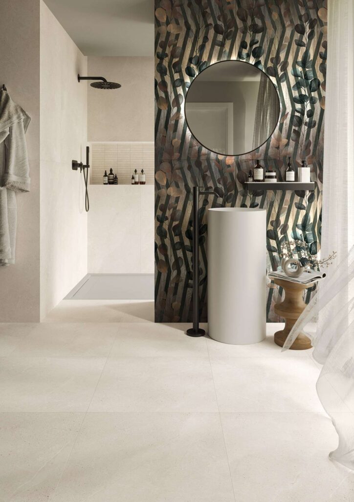 La collezione PALLADIO definisce questo elegante ambiente bagno di design, caratterizzato dal pavimento in formato 60x60 e dalle pareti della doccia in formato 60x120 nella delicata tonalità chiara Talco ad effetto pietra naturale. La parete principale è valorizzata dal suggestivo rivestimento Decoro Nuit in formato 60x120, impreziosito da un raffinato motivo botanico a foglie su righe scure verticali. Davanti al decoro si inseriscono un moderno lavabo a colonna bianco con miscelatore nero opaco a pavimento, un grande specchio circolare retroilluminato e un tavolino in legno, per un'atmosfera sofisticata e rilassante.