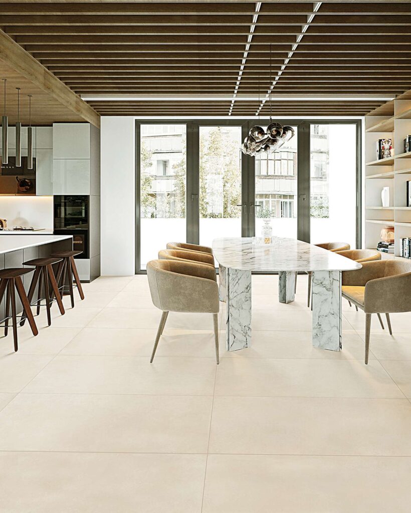 La collezione Proton Beige nel formato 60x120 cm definisce lo stile di una raffinata e luminosa sala da pranzo in un open space moderno. La pavimentazione in gres porcellanato beige chiaro, posata in modo regolare a correre, amplifica la luce naturale proveniente dalle ampie vetrate di sfondo. L'ambiente accogliente è arredato con un tavolo da pranzo con piano ed elementi in marmo venato, poltroncine coordinate in tessuto beige, una cucina di design con sgabelli in legno sulla sinistra e un caratteristico soffitto a doghe di legno che riscalda l'atmosfera.