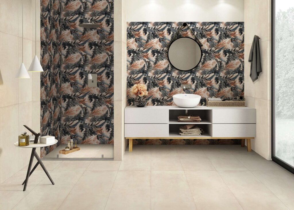 La collezione Proton Beige nel formato 60x120 riveste con eleganza questo bagno moderno, abbinando piastrelle a fondo neutro e caldo al decoro botanico Dec.Texdeco Warm con motivi a fogliame tropicale nei toni del verde e del ruggine. L'immagine mostra la posa a pavimento e a parete, con il decoro inserito sia all'interno della spaziosa doccia walk-in con vetro trasparente, sia sulla parete retrostante il mobile lavabo bianco. Un grande specchio circolare, un lavabo a bacinella e dettagli di design minimalisti completano l'atmosfera luminosa e raffinata dell'ambiente.