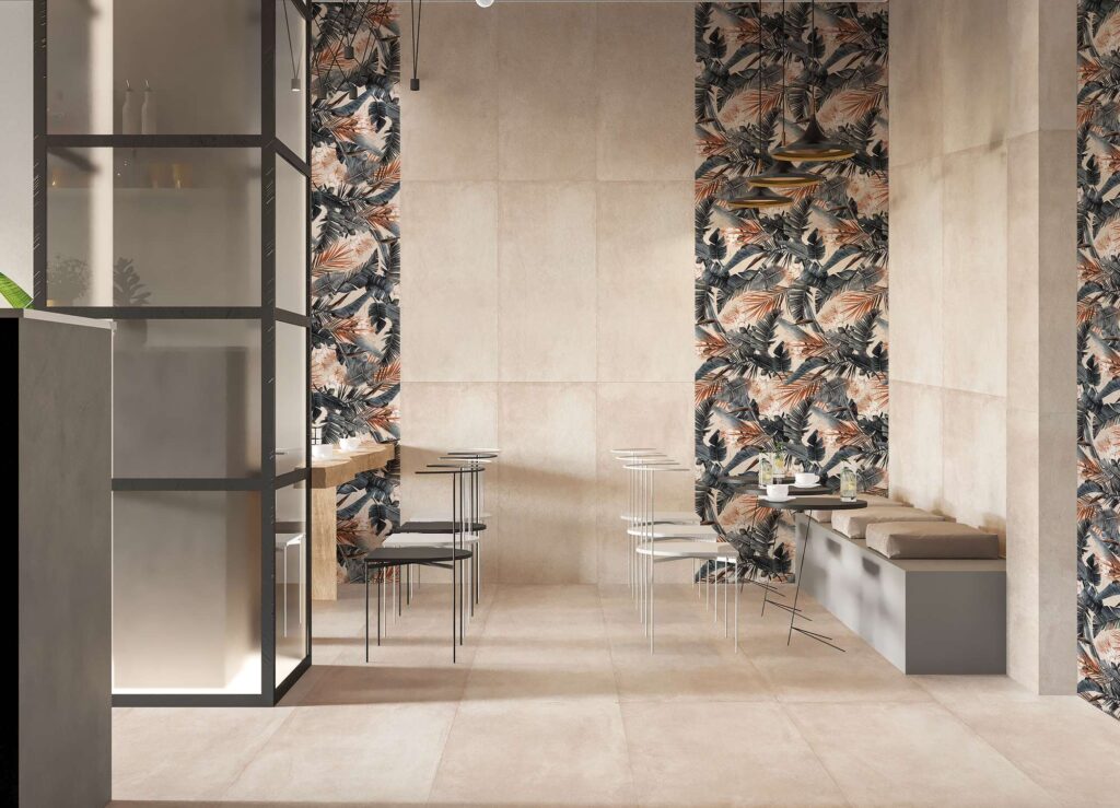 La collezione di piastrelle in gres porcellanato PROTON BEIGE, nel formato 60x120 cm, riveste con eleganza il pavimento e le pareti di questo moderno ristorante. La texture sobria e naturale nei toni del beige si sposa armoniosamente con le fasce decorative verticali a parete DEC.TEXDECO WARM, caratterizzate da un raffinato motivo botanico a fogliame nei toni caldi e naturali. L'ambiente, dall'atmosfera sofisticata e accogliente, è completato da arredi minimalisti come tavoli rotondi neri, sedie di design e una moderna panca con cuscini, valorizzati da una calda illuminazione sospesa.