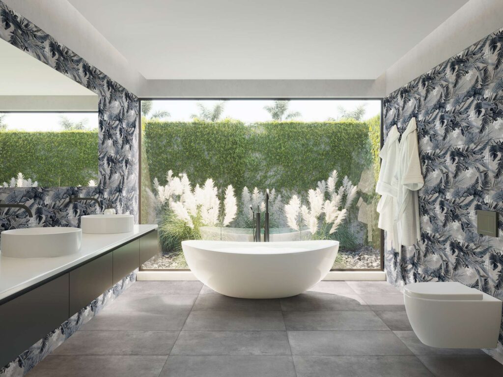 La collezione PROTON GRIS in gres porcellanato nel formato 60x120 definisce lo stile raffinato di questo bagno moderno e luminoso. Il pavimento è posato con grandi lastre grigie dall'effetto materico, mentre le pareti sono impreziosite dal rivestimento decorativo DEC.TEXDECO con un elegante motivo botanico a fogliame nei toni del grigio e del blu scuro. Al centro della stanza, una vasca da bagno freestanding bianca è posizionata davanti a un'ampia vetrata che si affaccia su un giardino verdeggiante, affiancata a sinistra da un mobile sospeso con doppio lavabo d'appoggio, creando un'atmosfera di benessere e design.