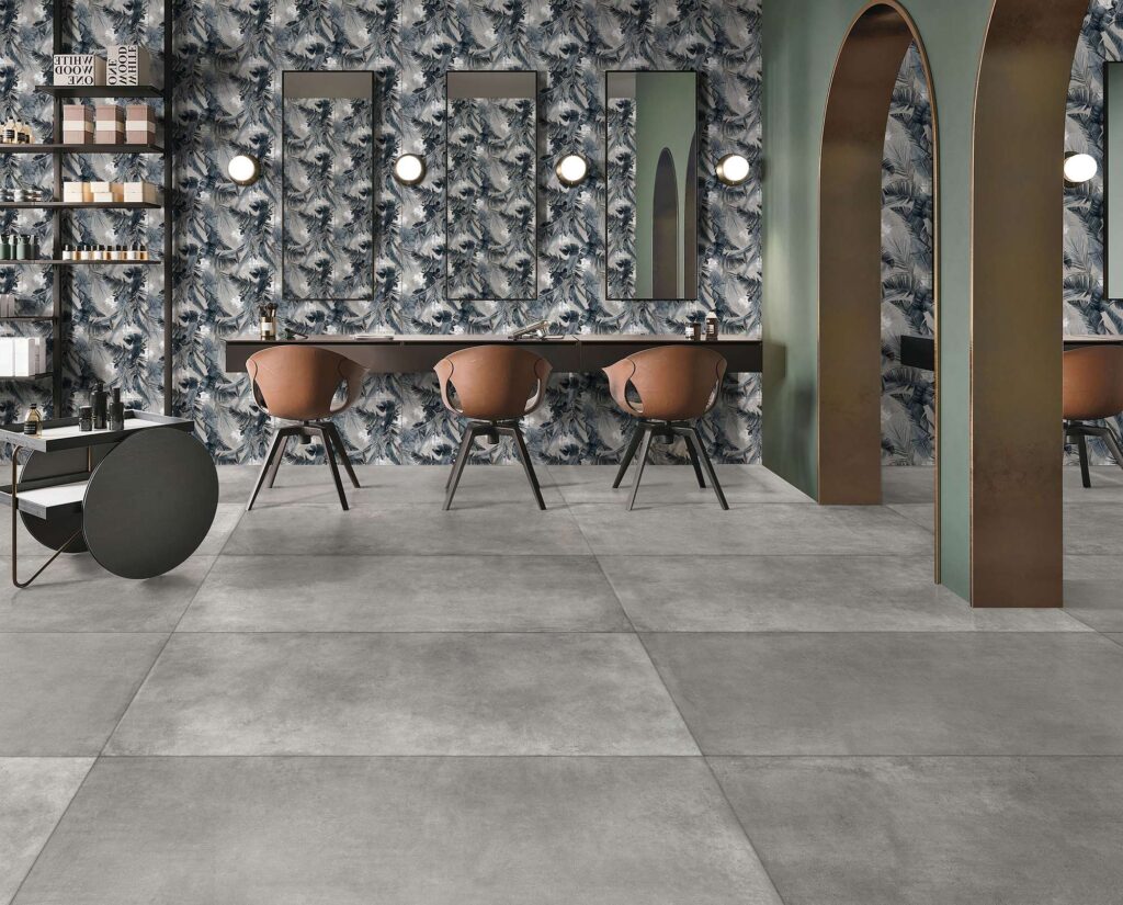 La collezione PROTON GRIS nel formato 60x120 definisce il pavimento di un moderno e sofisticato salone da parrucchiere con le sue piastrelle in gres porcellanato grigio effetto cemento. La parete principale è impreziosita dal decoro coordinato DEC.TEXTDECO COLD, caratterizzato da un motivo botanico nei toni freddi che fa da sfondo a specchi eleganti, poltroncine di design marroni e suggestivi archi decorativi color bronzo.