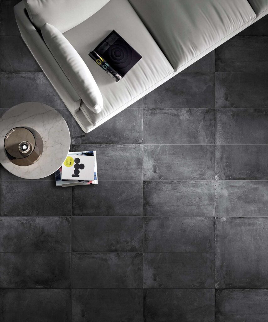 La collezione PROTON NOIR, nel formato 30x60 cm, veste il pavimento di questa raffinata zona living con piastrelle in gres porcellanato scuro dalla texture materica ed elegante. La vista dall'alto mostra la posa in linea delle piastrelle in tonalità nero antracite, accostate a elementi d'arredo minimalisti come un divano bianco di design, un tavolino rotondo in marmo chiaro e riviste d'arte, creando un contrasto cromatico sofisticato e contemporaneo.