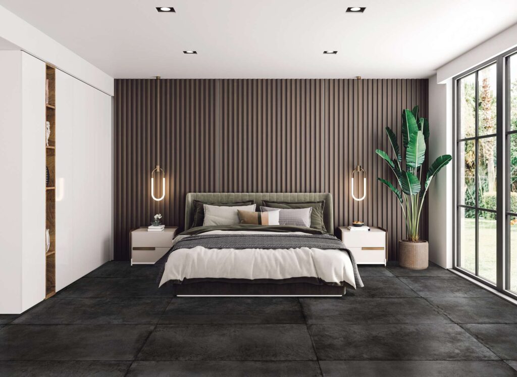La collezione PROTON NOIR nel formato 60x120 cm riveste il pavimento di questa camera da letto di design con lastre in gres porcellanato scuro effetto pietra, posate in modo regolare e continuo per un effetto moderno e d'impatto. L'ambiente, luminoso ed elegante, presenta un letto matrimoniale centrale accostato a una parete decorata con listelli di legno verticali scuri, illuminata da due lampade a sospensione sospese sopra i comodini bianchi. Sulla destra, una rigogliosa pianta tropicale in vaso e una grande vetrata affacciata sul verde esterno completano un'atmosfera lussuosa, accogliente e confortevole.