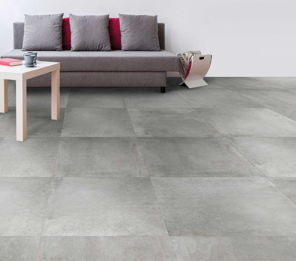 La collezione Proton, nel formato 60x60 e tonalità gris, riveste il pavimento di un moderno e luminoso soggiorno con piastrelle in gres porcellanato effetto cemento grigio. La posa lineare e regolare valorizza la superficie materica e contemporanea, inserendosi in un ambiente dal design minimalista arredato con un divano grigio arricchito da cuscini colorati, un tavolino da caffè chiaro e pareti bianche.