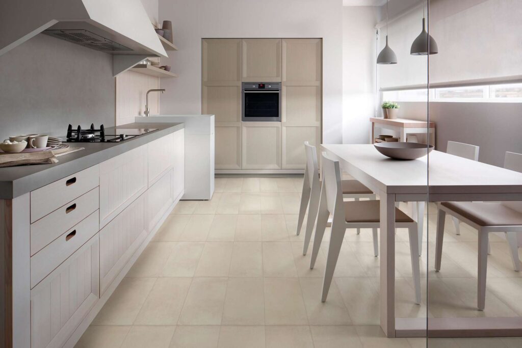 La collezione Proton in tonalità Beige, nel formato 30x60 cm, definisce il pavimento di una moderna cucina open space inondata di luce naturale. Le piastrelle in gres porcellanato, caratterizzate da una texture sobria e posate in modo regolare, creano un'atmosfera elegante e accogliente, armonizzandosi con i mobili bianchi della cucina, il top grigio e la zona pranzo adiacente con tavolo e sedie in tonalità coordinate.