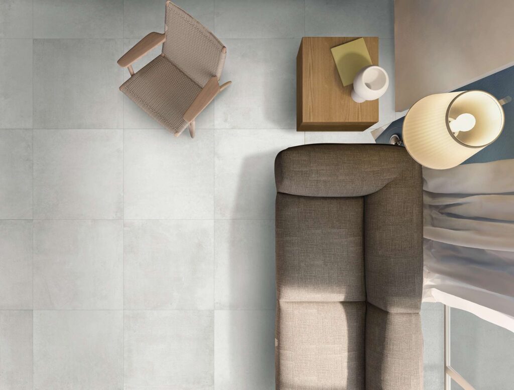 La collezione Proton Blanc, nel formato 60x60 cm, definisce con eleganza il pavimento di un soggiorno moderno in questa suggestiva vista dall'alto. Le piastrelle in ceramica nei toni del bianco e del grigio chiaro con effetto cemento sono posate in modo regolare, creando una base luminosa per un arredamento raffinato e minimalista: un divano in tessuto grigio, una poltroncina di design in legno e corda intrecciata, un tavolino in legno e una lampada da terra che diffonde una luce calda e rilassante.