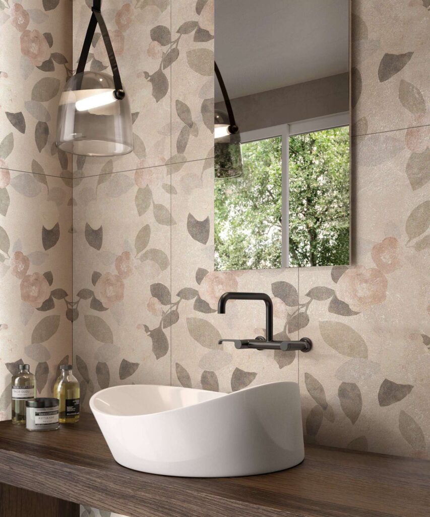 La collezione di piastrelle in ceramica QUINTESENCE DECORO PEONIES nel formato 60x120 cm decora con eleganza la parete di un raffinato bagno contemporaneo. Il rivestimento effetto carta da parati è caratterizzato da un delicato motivo floreale con peonie e foglie nei toni tenui del rosa cipria, del beige e del verde salvia. In primo piano, posato su un piano in legno scuro, risalta un moderno lavabo da appoggio bianco dalle linee sinuose e organiche, completato da una rubinetteria a parete nera opaca e da un elegante specchio rettangolare, creando un'atmosfera rilassante e di design.