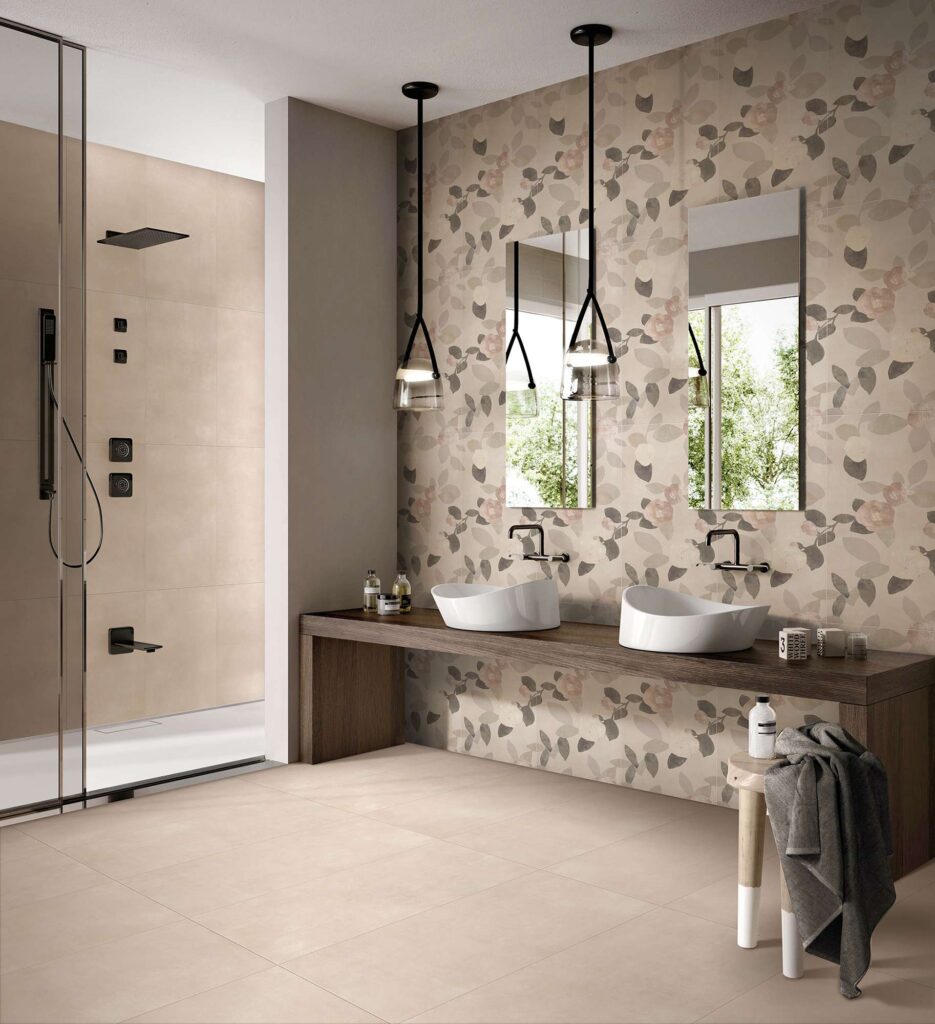 La collezione QUINTESSENCE in formato 60x120 cm nella sfumatura Beige riveste con eleganza il pavimento e la parete interna della doccia in questo bagno moderno e raffinato. La parete principale è valorizzata da un rivestimento decorativo a tema botanico, caratterizzato da un delicato motivo di foglie nei toni del grigio, beige e rosa antico. L'ambiente è completato da una consolle sospesa in legno scuro con due lavabi da appoggio bianchi, specchi verticali, lampade a sospensione nere e una doccia con eleganti dettagli in nero opaco.