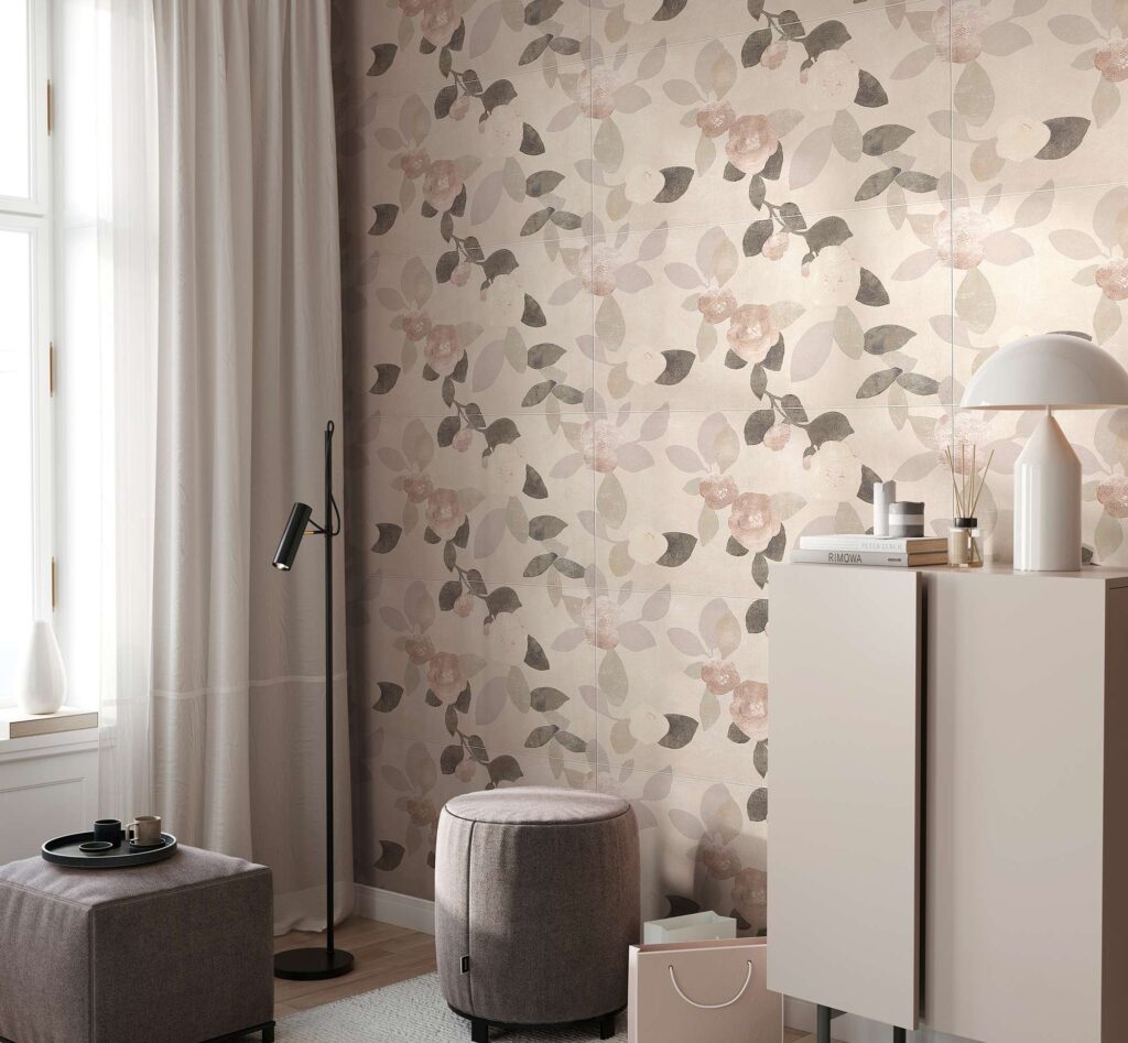 La collezione Quintessence Decoro, nel formato 60x120 cm, riveste con eleganza la parete di un moderno e luminoso soggiorno. Le piastrelle in ceramica presentano un raffinato motivo botanico con foglie e rose stilizzate nei toni del rosa cipria, grigio e delicati accenni metallici. L'ambiente, dall'atmosfera calda e accogliente, è completato da un arredamento minimalista composto da una madia chiara con lampada di design, due pouf in tessuto e una morbida tenda bianca che filtra la luce naturale proveniente dalla finestra.