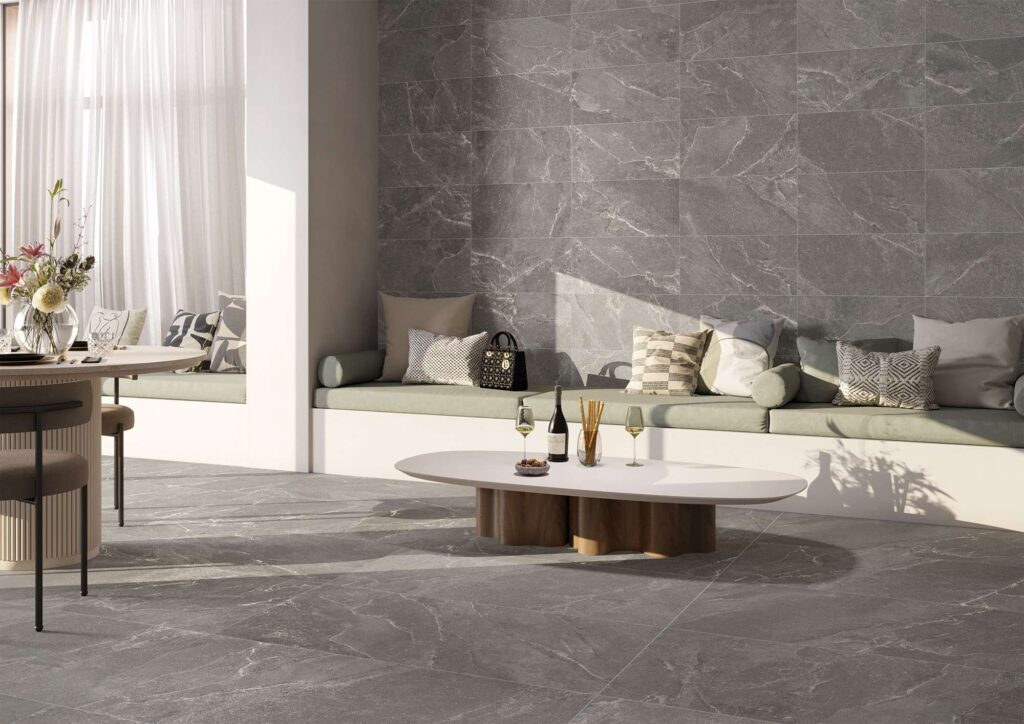 La collezione Reshape Charcoal valorizza questo luminoso spazio lounge dal design contemporaneo con le sue superfici in gres porcellanato effetto pietra grigio scuro. A pavimento spiccano le grandi lastre nel formato 60x120, mentre la parete retrostante è rivestita con il formato 30x60 posato orizzontalmente a correre. L'ambiente, elegante e accogliente, è arredato con una sofisticata panca a muro completata da morbidi cuscini decorativi in tonalità neutre e salvia, un tavolino da caffè basso e moderno, il tutto inondato da una calda luce naturale che esalta le venature naturali della ceramica.