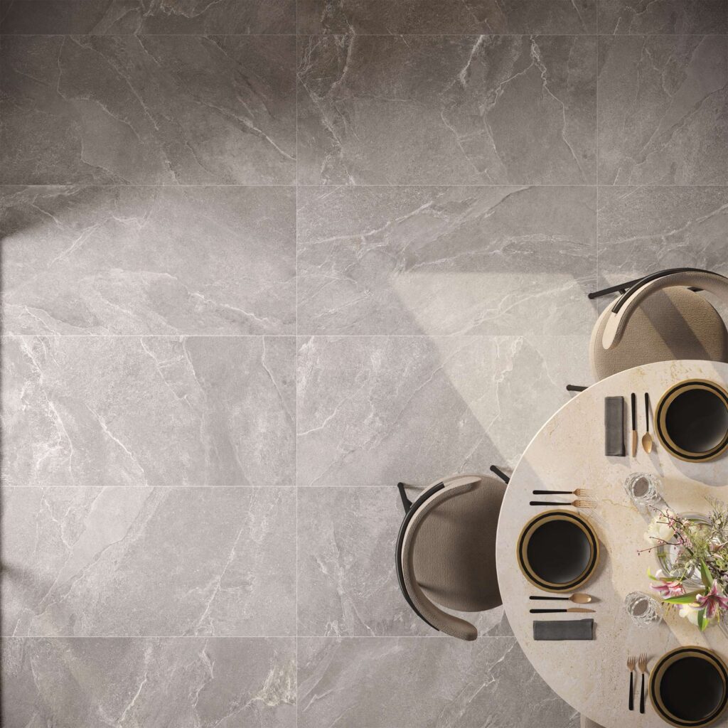 La collezione di piastrelle in gres porcellanato Reshape Charcoal, qui rappresentata nei formati 60x120 e 30x60, definisce con eleganza una raffinata zona pranzo. La veduta dall'alto mette in risalto il pavimento effetto pietra grigio scuro, caratterizzato da venature chiare e naturali. Accanto alla superficie ceramica, si svela parzialmente un moderno tavolo rotondo finemente apparecchiato con piatti scuri e dettagli dorati, circondato da sedie di design imbottite in tessuto tortora. Una calda luce naturale attraversa la scena, proiettando ombre geometriche che esaltano la texture materica e l'atmosfera accogliente e sofisticata dell'ambiente.