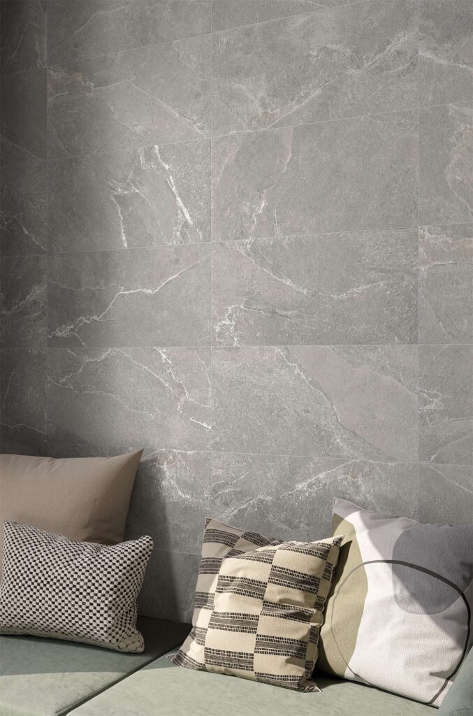 La collezione Reshape Charcoal, disponibile nei formati 60x120 e 30x60, decora la parete di un raffinato spazio d'attesa con piastrelle in gres porcellanato effetto pietra grigia arricchite da venature chiare e naturali. In primo piano, una seduta moderna con cuscini decorativi dalle trame geometriche e tonalità neutre è lambita da una calda luce diagonale che esalta la texture materica del rivestimento a parete.