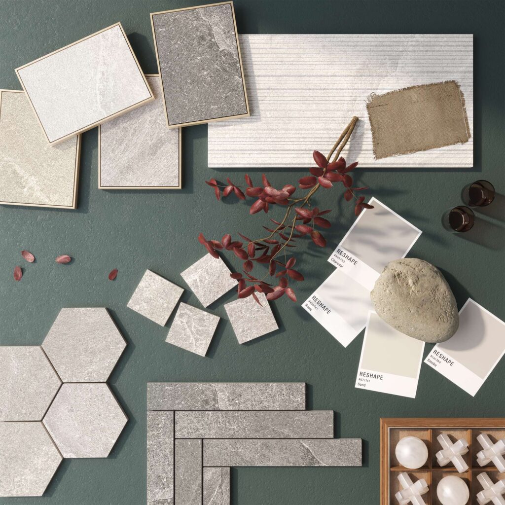 La collezione RESHAPE è protagonista di un raffinato moodboard flat lay su uno sfondo verde scuro materico, che mette in mostra la versatilità delle sue superfici ceramiche effetto pietra. L'immagine presenta diversi formati della collezione, tra cui piastrelle esagonali, listelli rettangolari posati a spina di pesce, piccoli mosaici quadrati e grandi lastre con texture rigata. I cartoncini campionari evidenziano le varianti colore Charcoal, Snow, Sand e Smoke. L'atmosfera elegante e naturale è completata da elementi decorativi come un ramo con foglie ramate, un ciottolo di fiume, un frammento di tessuto in juta e un vassoio in legno, evocando un design d'interni armonioso e sofisticato.