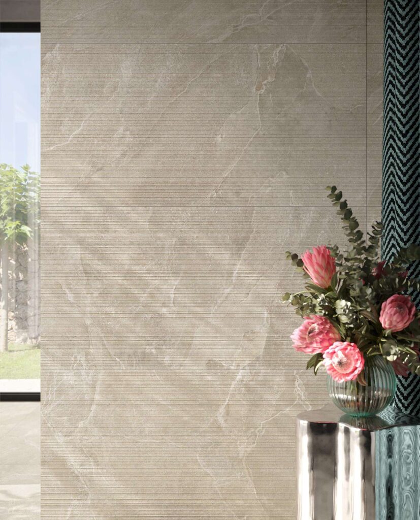 La collezione Reshape Sand nel formato 60x120 Lines riveste la parete di un raffinato bagno contemporaneo. L'immagine mostra le piastrelle ceramiche color sabbia caratterizzate da una texture tridimensionale a righe orizzontali e delicate venature effetto pietra. Sulla destra, un vaso di vetro scanalato con fiori rosa e foglie di eucalipto poggia su un moderno tavolino metallico lucido, accanto a un elemento decorativo con motivo chevron geometrico. Sulla sinistra, la luce naturale entra da una vetrata aperta sul giardino esterno, valorizzando la posa a parete e l'atmosfera calda e accogliente dell'ambiente.