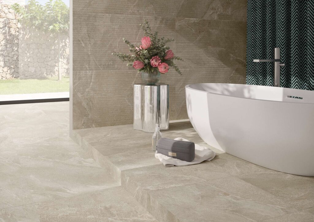 La collezione RESHAPE SAND 60x120 in gres porcellanato effetto pietra definisce lo stile di un bagno moderno e luminoso. La pavimentazione color sabbia a grandi lastre si sviluppa su un elegante piano rialzato, coordinandosi con il rivestimento a parete caratterizzato dalla raffinata texture a righe orizzontali "Lines". L'ambiente è completato da una vasca freestanding bianca di design, un tavolino metallico riflettente con un vaso di fiori rosa e un'ampia vetrata con vista su un giardino esterno che esalta la naturalezza e la luminosità del design.