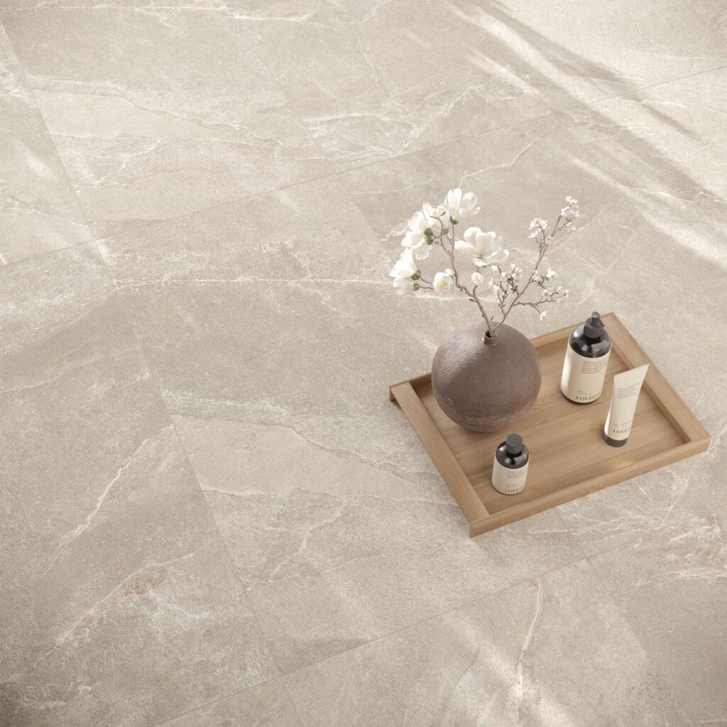 La collezione Reshape Sand nel formato 60x120 cm riveste il pavimento con eleganti piastrelle in gres porcellanato effetto pietra dalle calde tonalità sabbia, arricchite da delicate venature chiare. Sulla superficie opaca è adagiato un vassoio in legno minimalista che ospita un vaso sferico con rami fioriti bianchi e flaconi di cosmetici, evocando un'atmosfera di puro benessere e relax. Una calda luce naturale attraversa la stanza, creando morbide ombre diagonali che esaltano la texture materica e il design raffinato e contemporaneo dell'ambiente.