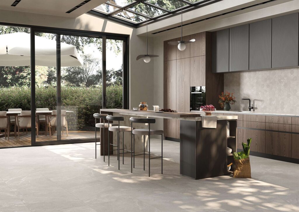 La collezione Reshape Smoke nel formato 60x120 cm definisce lo stile di una cucina moderna e luminosa di alto design attraverso un pavimento in gres porcellanato grigio effetto pietra. L'ambiente, caratterizzato da un'elegante isola centrale con sgabelli coordinati e caldi arredi in legno, si apre verso l'esterno grazie a un'ampia vetrata e a un lucernario a soffitto. La luce naturale filtra nella stanza valorizzando la posa lineare e la texture sofisticata e naturale delle grandi lastre ceramiche.