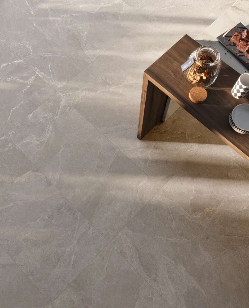 La collezione Reshape nella tonalità Smoke, formato 60x120, riveste il pavimento di un'elegante cucina con piastrelle in gres porcellanato effetto pietra grigia posate in diagonale. La superficie materica, arricchita da delicate venature chiare, è lambita da una calda luce naturale che crea morbidi giochi di luce e ombra. In alto a destra, l'angolo di un tavolo in legno scuro con un barattolo di biscotti e tazze da colazione completa la scena, evocando un'atmosfera accogliente, intima e di design.