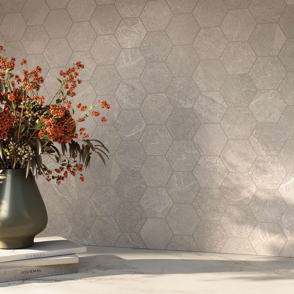 La collezione Reshape Smoke nel formato 60x120 cm definisce lo stile di questa cucina raffinata attraverso un elegante rivestimento a mosaico esagonale con texture effetto pietra nei toni del grigio fumo. In primo piano, su un piano di lavoro in marmo chiaro accarezzato da una calda luce naturale, si trova un vaso scuro con rami decorativi e bacche arancioni appoggiato su libri di design, creando un'atmosfera accogliente e contemporanea.