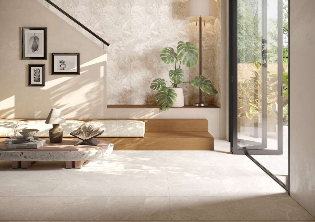 La collezione Reshape, qui nella tonalità Snow in formato 60x60 cm, definisce la pavimentazione di un elegante e luminoso ambiente living dal design contemporaneo. Le piastrelle in gres porcellanato chiaro effetto pietra, posate con fuga minima, riflettono la calda luce naturale che entra da un'ampia vetrata scorrevole. L'atmosfera rilassante e d'ispirazione naturale è completata da arredi minimalisti in legno, una rigogliosa pianta di monstera e una parete di fondo decorata con delicati motivi botanici coordinati nei toni del beige.