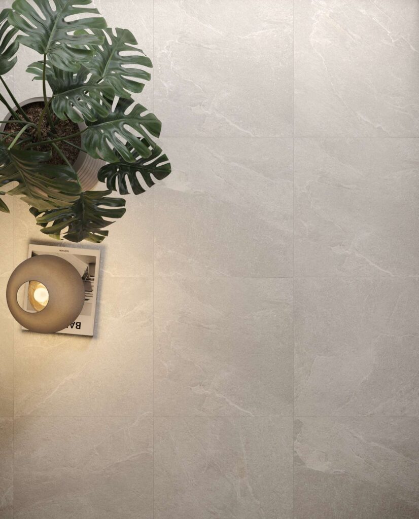 La collezione Reshape nel colore Snow, qui nel formato 60x60 cm, riveste il pavimento di un elegante ambiente living con un gres porcellanato effetto pietra dalle tonalità chiare e naturali. L'inquadratura dall'alto evidenzia la posa regolare e la texture materica delle piastrelle, arricchite nell'angolo sinistro da raffinati dettagli d'arredo: una rigogliosa pianta di monstera in vaso, un libro di design e una lampada sferica accesa che diffonde una luce calda e accogliente sulla superficie.