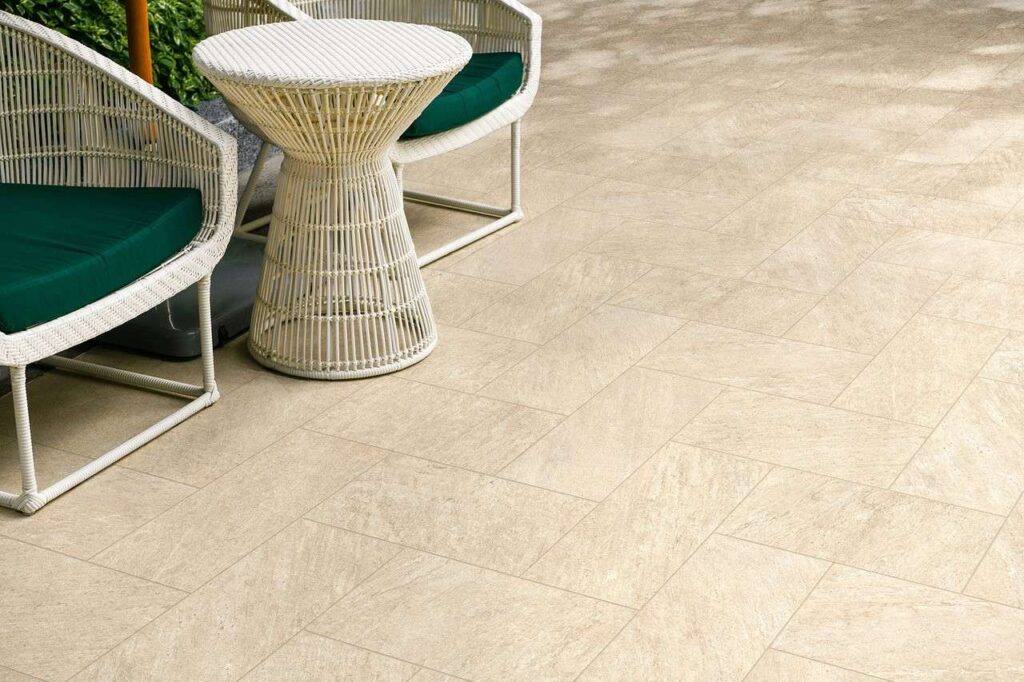 La collezione Seattle Beige, qui nel formato 20x40 cm ideale per l'outdoor, riveste con eleganza il pavimento di un patio esterno grazie a piastrelle in gres porcellanato effetto pietra naturale posate con una raffinata disposizione a spina di pesce. Sulla pavimentazione texturizzata dalle calde tonalità del beige è posizionato un salottino da giardino composto da due poltroncine e un tavolino in vimini intrecciato bianco con cuscini verde bosco, creando un'atmosfera accogliente, luminosa e contemporanea.