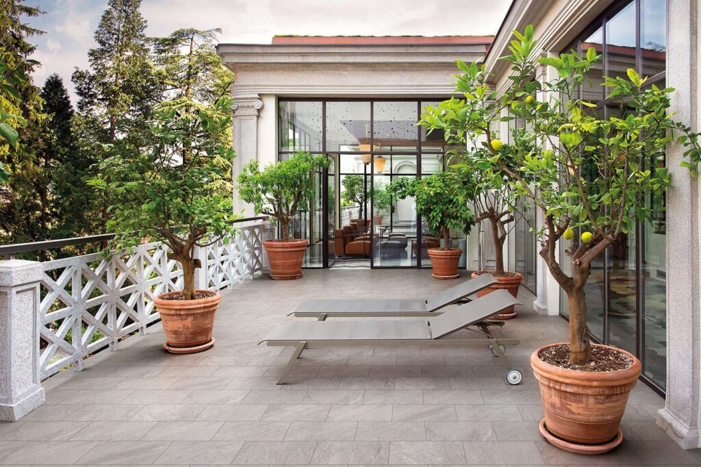 Collezione Seattle Grigio nel formato 20x40,3 per esterni, utilizzata per la pavimentazione con posa a correre di un lussuoso terrazzo panoramico. Il gres porcellanato effetto pietra grigia dona eleganza e continuità visiva all'ambiente outdoor, impreziosito da grandi vasi in terracotta con rigogliosi alberi di limone, lettini prendisole moderni e ampie vetrate di una villa signorile.