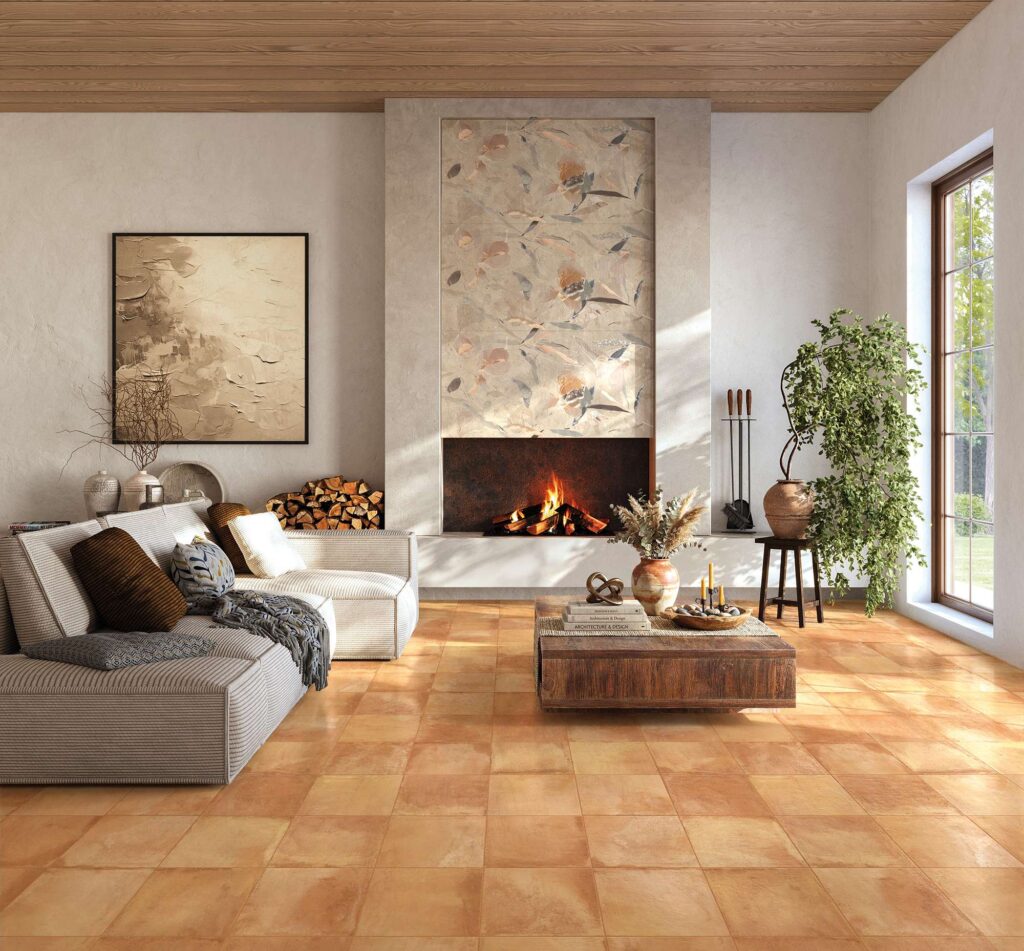 La collezione TRADIZIONE ROSATO TOSCANO nel formato 30x30 arreda con calore e raffinatezza un luminoso soggiorno (living), mostrando un pavimento in ceramica effetto cotto dalle sfumature calde e rosate posato in modo regolare. Sullo sfondo, incorniciato sopra un moderno caminetto acceso, risalta il decoro a parete AQUARELLE con delicati motivi artistici d'ispirazione naturale. L'ambiente, caratterizzato da un'atmosfera accogliente e sofisticata, è completato da un divano di design nei toni del grigio, un tavolino in legno rustico e piante ornamentali, valorizzati dalla luce naturale che entra da un'ampia finestra.