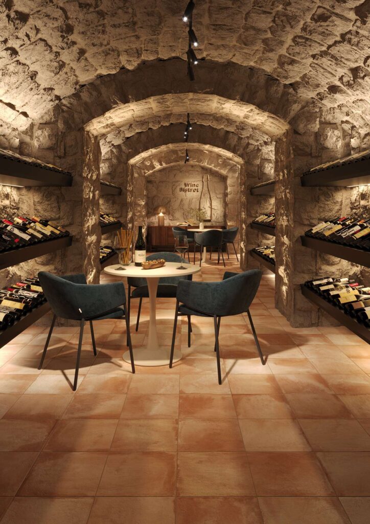 Collezione TRADIZIONE: pavimento effetto cotto nella variante Cotto Fiorentino formato 30x30, caratterizzato da sfumature calde e tradizionali posate con fuga regolare. L'immagine mostra una suggestiva cantina di degustazione (wine bistrot) dall'atmosfera calda e accogliente, definita da pareti e soffitti a volta in pietra grezza. L'ambiente è arredato con tavoli rotondi bianchi, eleganti poltroncine in tessuto blu scuro e scaffali retroilluminati colmi di bottiglie di vino, che esaltano il carattere rustico e raffinato del pavimento.