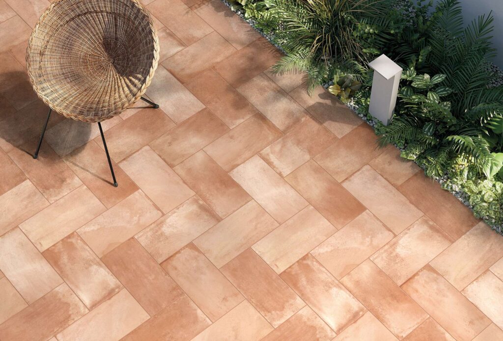 La collezione Tradizione presenta la piastrella in gres porcellanato effetto cotto nella variante Cotto Fiorentino, formato 20x40,3 cm, ideale per la pavimentazione di ambienti outdoor. L'immagine mostra una posa a spina di pesce dall'alto, caratterizzata dalle calde sfumature della terra cotta che creano un'atmosfera accogliente e solare. Nell'angolo sinistro è visibile una sedia di design in midollino intrecciato, mentre il lato destro è arricchito da un'aiuola con piante verdi rigogliose e un moderno lampioncino da esterno.