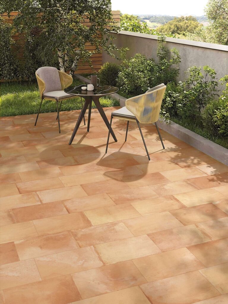 La collezione TRADIZIONE, nella variante colore Rosato Toscano in formato 20x40,3, definisce lo stile di questo raffinato ambiente outdoor dal carattere caldo e accogliente. L'immagine mostra una pavimentazione per esterni effetto cotto con posa sfalsata, caratterizzata da calde sfumature rosate, aranciate e sabbia che catturano la luce naturale. Sul patio, circondato da un giardino curato con piante verdi e una vista panoramica sulle colline, è allestita un'elegante area relax con un tavolino rotondo scuro e due sedie di design in corda intrecciata, creando un'atmosfera solare e di quiete estiva.