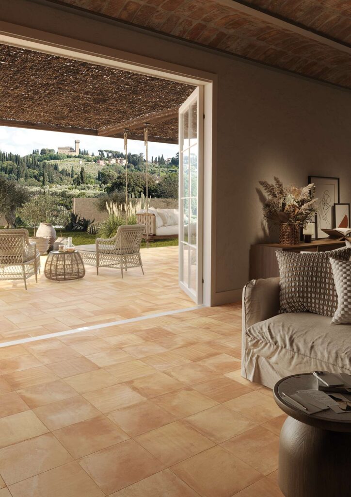 Collezione TRADIZIONE: pavimentazione in gres porcellanato effetto cotto nella calda tonalità Rosato Toscano, posata nei formati 30x30 e 20x40. L'immagine mostra una raffinata continuità visiva tra un elegante ambiente living interno, caratterizzato da soffitto con voltine in mattoni e arredi accoglienti, e un patio esterno coperto da un pergolato e arredato con poltrone in vimini. La posa armonica del pavimento valorizza il passaggio tra indoor e outdoor, incorniciando un suggestivo panorama collinare toscano sullo sfondo.
