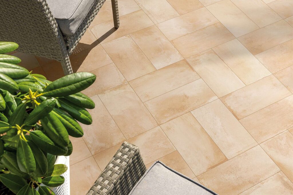 La collezione Tradizione, nella calda tonalità Umbro Paglierino in formato 20x40,3 cm, valorizza questo ambiente outdoor con un'elegante posa a spina di pesce. L'immagine mostra il pavimento per esterni in gres porcellanato effetto cotto, caratterizzato da sfumature naturali e sabbiose, inserito in un patio soleggiato e accogliente, arredato con poltrone in rattan grigio complete di cuscini e una pianta verde rigogliosa in primo piano.