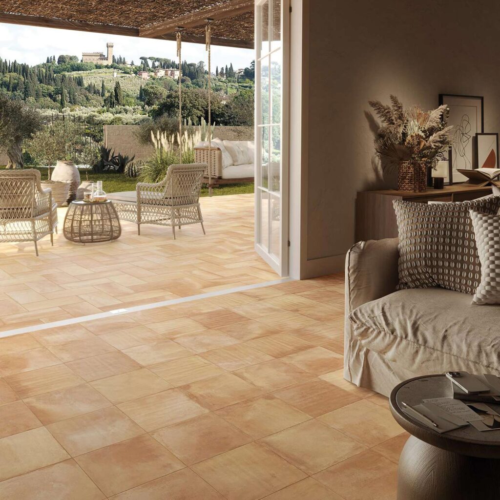 Collezione TRADIZIONE: pavimentazione in gres porcellanato effetto cotto nella calda tonalità Rosato Toscano, posata nei formati 30x30 e 20x40. L'immagine mostra una raffinata continuità visiva tra un elegante ambiente living interno, caratterizzato da soffitto con voltine in mattoni e arredi accoglienti, e un patio esterno coperto da un pergolato e arredato con poltrone in vimini. La posa armonica del pavimento valorizza il passaggio tra indoor e outdoor, incorniciando un suggestivo panorama collinare toscano sullo sfondo.