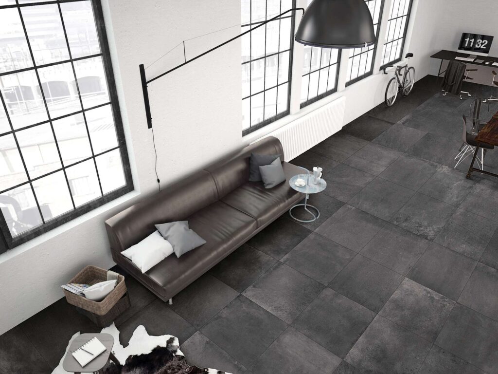 La collezione UNIKA CARBON nel formato 60x60 cm riveste il pavimento di un ampio e luminoso living in stile loft industriale. La vista dall'alto mostra la posa regolare delle piastrelle in gres porcellanato color grigio scuro antracite con effetto materico, accostate a grandi finestre stile officina, pareti bianche, un divano in pelle scura e un angolo studio sullo sfondo.