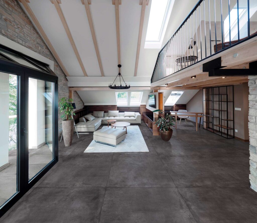 Pavimento in gres porcellanato della collezione Unika Carbon nel formato 80x80 cm, posato all'interno di un ampio e luminoso living moderno caratterizzato da un soffitto con travi a vista e un soppalco in legno. Le piastrelle grigio scuro effetto pietra, con posa ortogonale, si estendono con eleganza in tutto l'ambiente, dialogando con gli arredi contemporanei come il divano ad angolo chiaro, il tappeto geometrico e le grandi vetrate che si aprono sull'esterno creando continuità visiva.