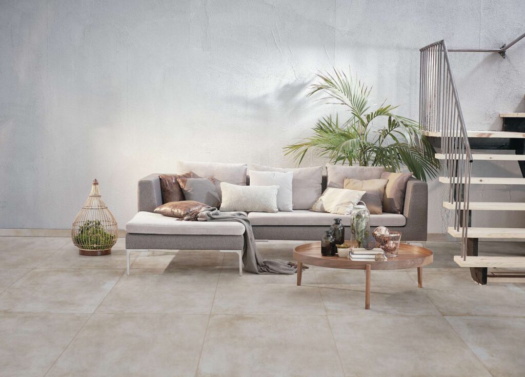 La collezione Unika Sand nel formato 80x80 riveste il pavimento di un moderno e luminoso ambiente living. Le piastrelle in gres porcellanato color sabbia, con posa ortogonale e fughe minimali, donano ampiezza e continuità visiva allo spazio. L'arredamento, elegante e dal design contemporaneo, comprende un divano grigio arricchito da cuscini in tonalità calde e naturali, un tavolino basso in legno rotondo con dettagli decorativi, una pianta tropicale e una scala a giorno sullo sfondo, il tutto incorniciato da una parete materica dallo stile minimalista.