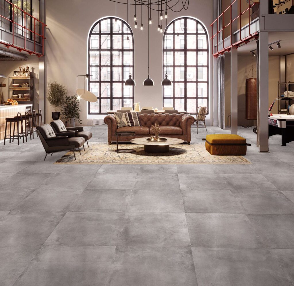 La collezione Unika, nella sfumatura grigia Smoke in formato 60x60 cm, definisce con eleganza il pavimento di un ampio e prestigioso soggiorno in stile loft industriale. Il gres porcellanato effetto pietra, posato in una griglia regolare e uniforme, riflette la luce naturale proveniente dalle imponenti finestre ad arco a doppia altezza. L'ambiente è valorizzato da arredi di classe, tra cui un classico divano Chesterfield in pelle marrone posizionato su un tappeto decorato, poltrone moderne e calde luci a sospensione, per un'atmosfera sofisticata, accogliente e dal design contemporaneo.