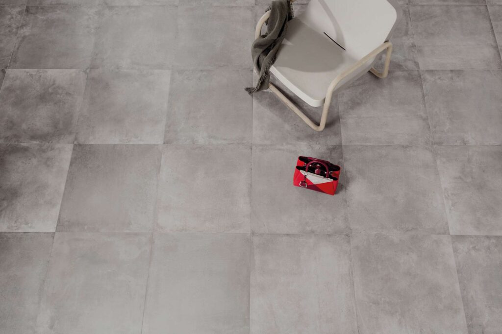 La collezione Unika, nella tonalità Smoke e formato 60x60 cm, si esprime in un elegante pavimento in gres porcellanato grigio effetto cemento dalle sfumature morbide e contemporanee. La vista dall'alto cattura la texture materica e la posa regolare delle piastrelle, accostate a una sedia di design minimalista con un telo grigio e a una vivace borsa rossa che risalta sul pavimento creando un raffinato contrasto cromatico.