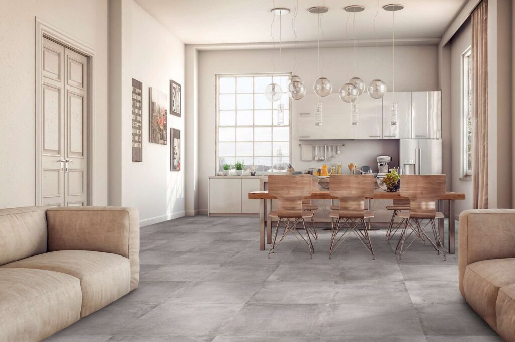 La collezione di piastrelle in gres porcellanato UNIKA nella variante colore SMOKE, formato 60x60 cm, definisce con eleganza il pavimento di una luminosa cucina con zona giorno open space. La posa lineare delle grandi piastrelle grigie effetto cemento si integra armoniosamente in un ambiente moderno e arioso, caratterizzato da un tavolo da pranzo in legno con sedie di design, lampade a sospensione sferiche in vetro, arredi da cucina minimalisti bianchi e accoglienti divani beige in primo piano.