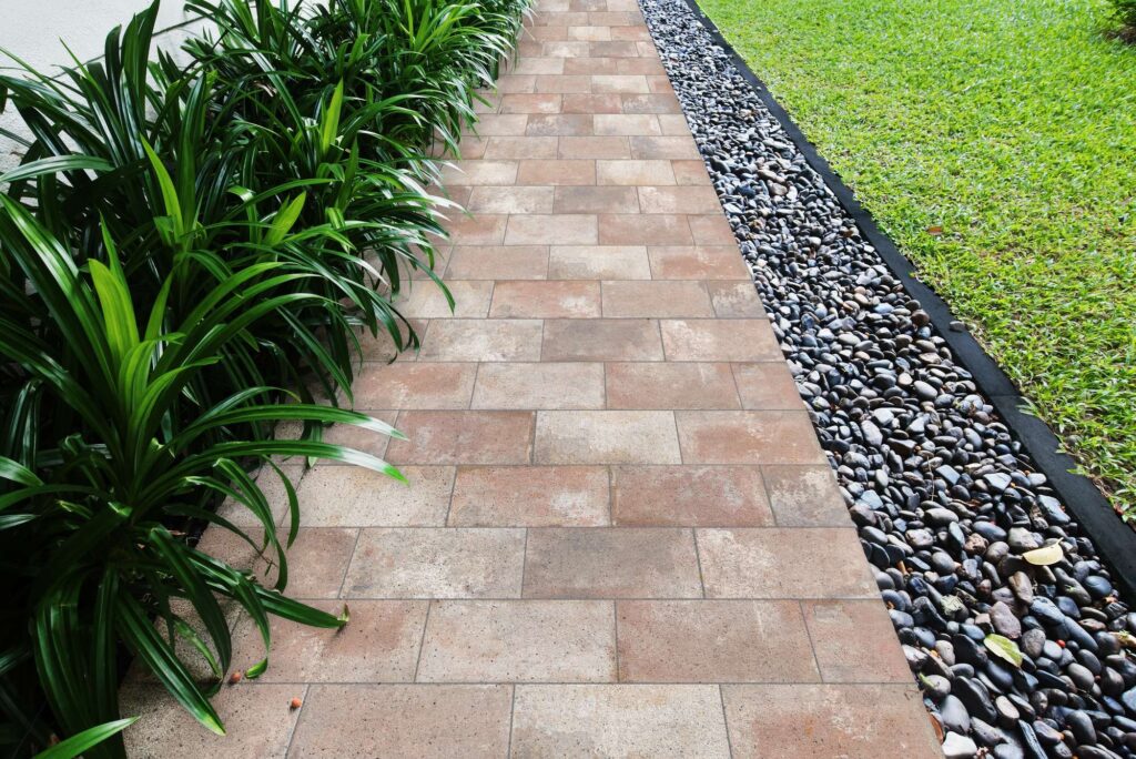 La collezione VENTIQUARANTA COTTO MIX in gres porcellanato effetto cotto definisce un elegante camminamento esterno con piastrelle nel classico formato rettangolare 20x40 cm. La posa sfalsata a correre valorizza le calde sfumature della terra e del terracotta che caratterizzano la finitura, creando un marciapiede armonioso circondato da una fitta vegetazione verde sulla sinistra e da una fascia decorativa di ciottoli di fiume scuri e prato erboso sulla destra.