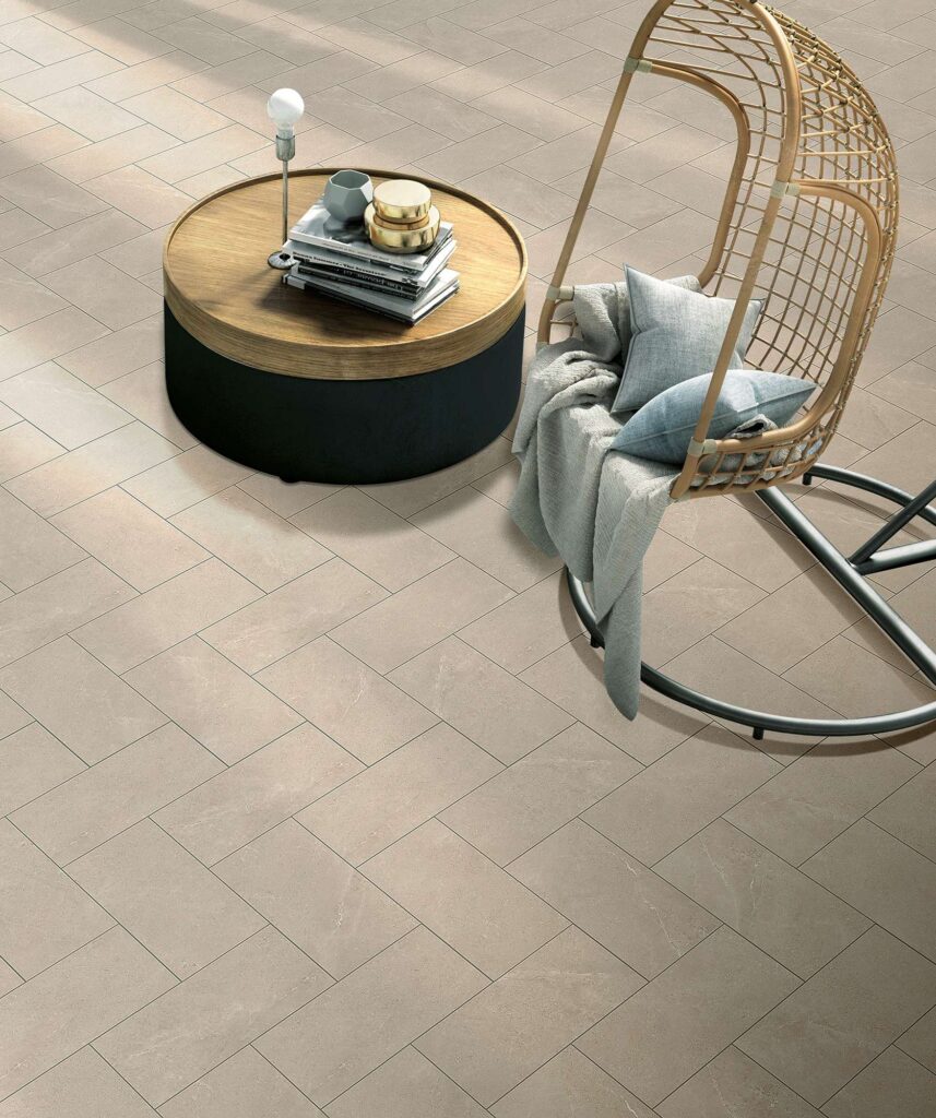 Pavimento per esterni della collezione VENTIQUARANTA in gres porcellanato color Blend Tan, nel formato 20x40 cm con posa diagonale a spina di pesce. L'immagine mostra un raffinato ambiente outdoor inondato da una calda luce naturale, arredato con un'elegante sedia sospesa in rattan con morbidi cuscini e un plaid, affiancata da un tavolino basso rotondo in legno e metallo con libri e oggetti di design che valorizzano la texture naturale della ceramica.