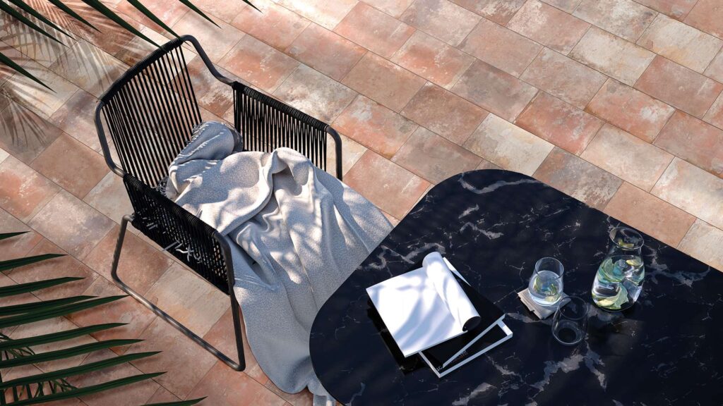 Collezione VENTIQUARANTA in variante Cotto: pavimento per esterni in gres porcellanato nel formato 20x40 cm con posa sfalsata a correre. L'inquadratura dall'alto mostra una raffinata zona relax outdoor, dove le piastrelle dalle calde sfumature effetto cotto fanno da sfondo a una poltrona di design in corda nera con una morbida coperta grigia e a un tavolino in marmo scuro con venature bianche, il tutto impreziosito da suggestivi giochi di luci e ombre proiettati da foglie di palma.