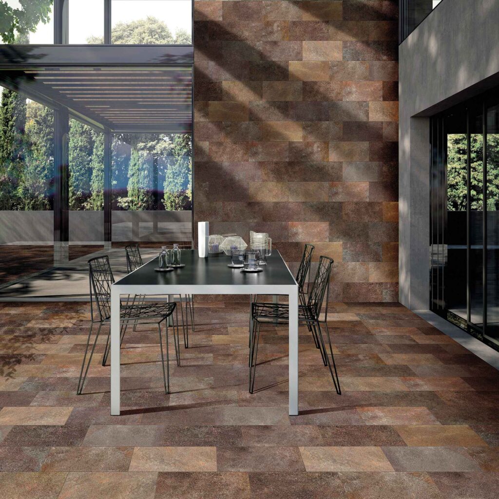 La collezione VENTIQUARANTA, nel formato 20x40 e nella calda tonalità Porfido Brown, caratterizza questo elegante ambiente outdoor con piastrelle in gres porcellanato effetto pietra naturale. La posa sfalsata (a correre) riveste con armonia sia il pavimento che la parete di fondo, esaltando le ricche sfumature marroni, bronzee e ramate delle superfici. Al centro del patio di una villa contemporanea, un moderno tavolo da pranzo con sedie geometriche in metallo nero completa l'allestimento, immerso in un'atmosfera lussuosa e accogliente definita da suggestivi giochi di luce naturale e ombre.