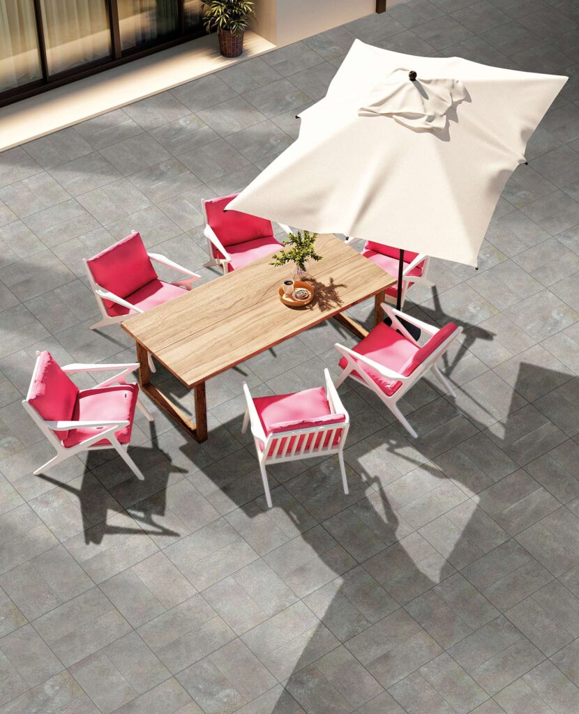 La collezione VENTIQUARANTA presenta la pavimentazione per esterni Porfido Grey nel formato 20x40 cm, un gres porcellanato effetto pietra ideale per l'outdoor. L'immagine mostra una splendida terrazza soleggiata caratterizzata dalla posa sfalsata delle piastrelle grigie, arredata con un tavolo da pranzo in legno, sedie bianche di design impreziosite da vivaci cuscini rosa acceso e un grande ombrellone color crema che proietta un'elegante ombra geometrica sullo spazio aperto.