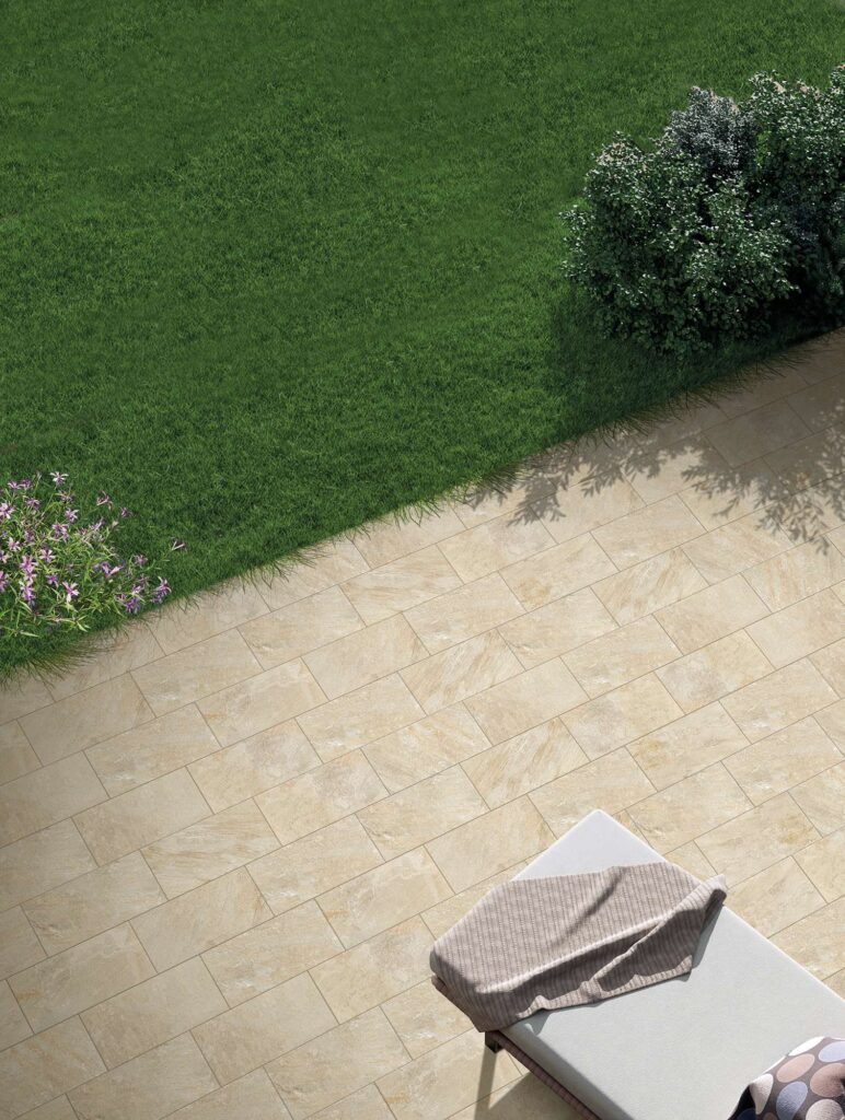Collezione VENTIQUARANTA: pavimentazione per esterni realizzata con piastrelle Rockline Beige nel formato 20x40 cm. L'immagine mostra una raffinata posa sfalsata (a correre) di ceramiche effetto pietra che delimitano un prato verde curato, arricchito da cespugli e delicati fiori viola. In basso a destra, la presenza di un elegante lettino prendisole con un telo morbido evoca un'atmosfera di relax all'aria aperta, illuminata da una calda luce naturale.
