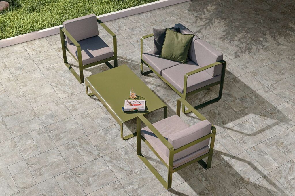 La collezione VENTIQUARANTA, nella variante Rockline Grey in formato 20x40, valorizza questo spazio outdoor con una pavimentazione in gres porcellanato effetto pietra grigia ricca di sfumature naturali. Le piastrelle, posate con un disegno geometrico alternato, ospitano un elegante salotto da giardino composto da un divano e due poltrone in metallo verde oliva con cuscini chiari, disposti attorno a un tavolino basso coordinato. L'atmosfera è rilassante e contemporanea, arricchita dalle morbide ombre di alberi che si posano sul pavimento e da un accenno di prato verde curato sullo sfondo.