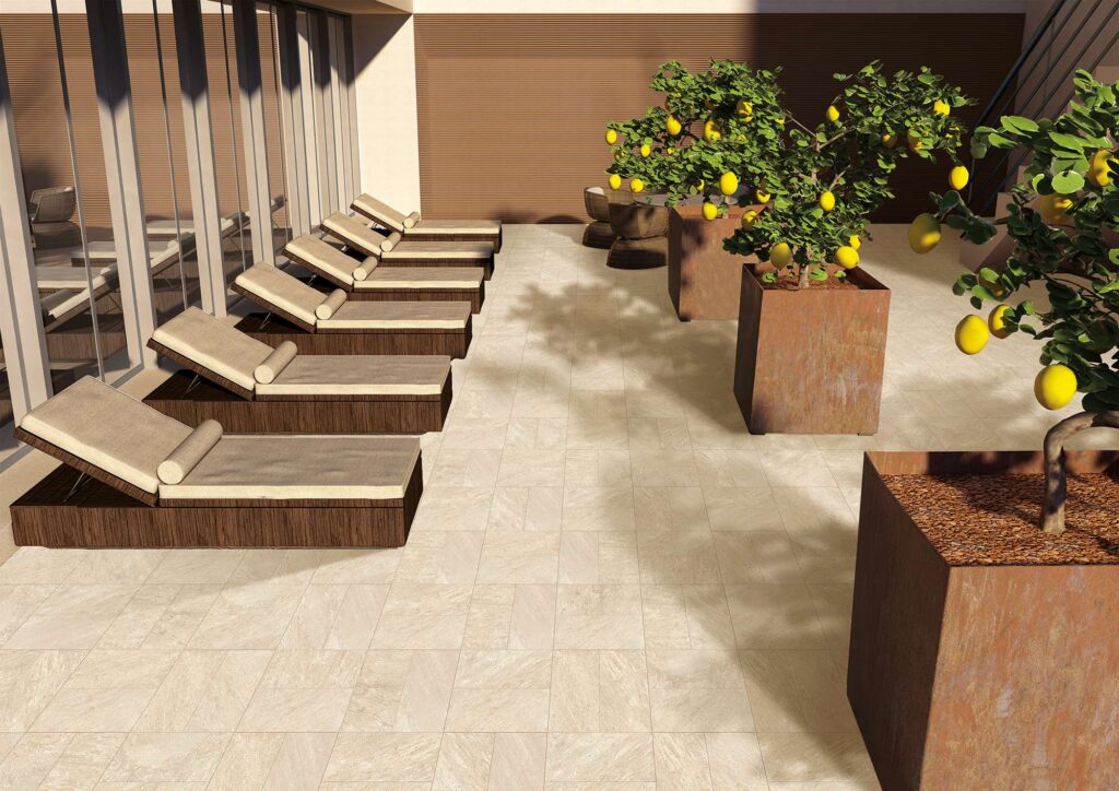 La collezione VENTIQUARANTA, nella raffinata tonalità Seattle Beige e formato 20x40, valorizza questo spazioso terrazzo outdoor dal design moderno e solare. La pavimentazione esterna, caratterizzata da piastrelle effetto pietra posate a moduli geometrici alternati, accoglie sul lato sinistro una fila di eleganti lettini prendisole in legno con cuscini beige, disposti lungo una grande vetrata. Sulla destra, grandi fioriere squadrate in corten ospitano rigogliosi alberi di limone carichi di frutti, che proiettano ombre nitide sulla superficie ceramica, ricreando un'atmosfera calda, lussuosa e tipicamente mediterranea.