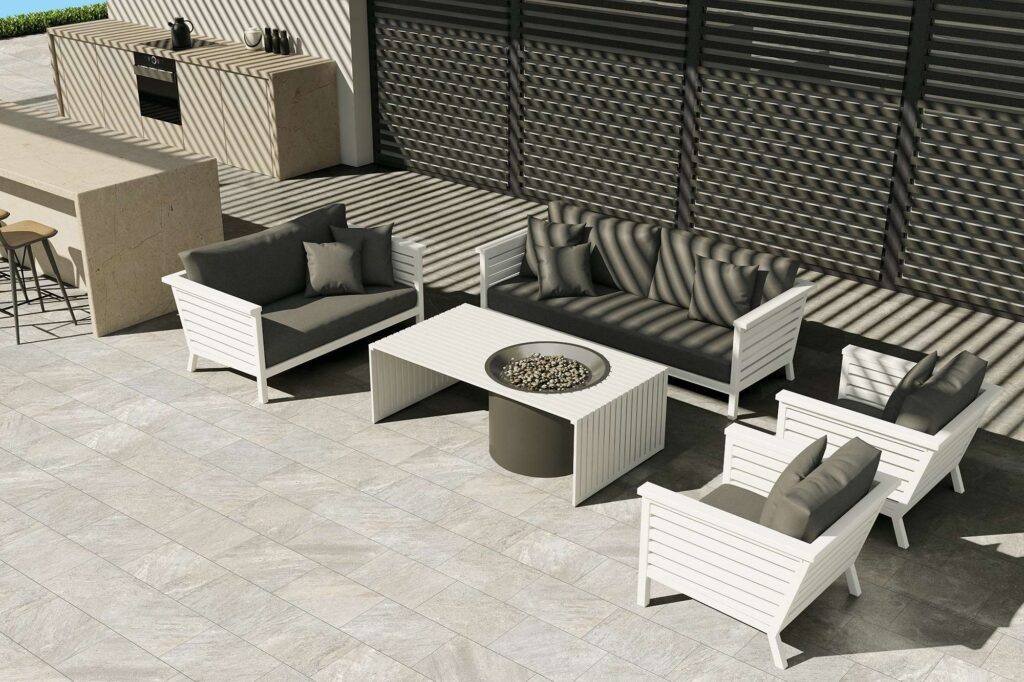 La collezione VENTIQUARANTA, nella raffinata tonalità Seattle Grey e nel formato 20x40, definisce lo stile di questo elegante spazio outdoor. La pavimentazione in gres porcellanato effetto pietra, caratterizzata da una posa a correre sfalsata, ospita un lussuoso salotto da giardino con divani e poltrone bianchi dai cuscini scuri, disposti attorno a un tavolino con braciere centrale. Sullo sfondo si scorge una cucina da esterno dal design minimale, mentre una luce solare radente proietta un suggestivo gioco di ombre geometriche sulla superficie, esaltando l'atmosfera sofisticata e accogliente del patio.